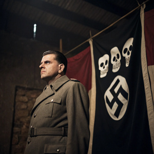Josef Mengele, standing in front of a dark, ominous banner with skull imagery. Dimly lit indoor setting.