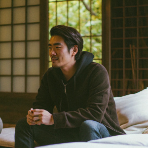 Takeshi Kaneshiro, in a candid photo, sitting on a minimalist sofa. Modern art piece in background. Dressed in a casual black hoodie and dark jeans.