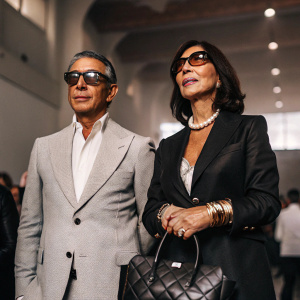 Mark Mandel and Emilia Pirro Mandel attending a gala event. Mark in a gray suit with a shirt and attached sunglasses. Emilia in a black blazer with pearl necklace and gold bracelet. Holding structured black leather handbag. Softly lit indoor venue with crowd.