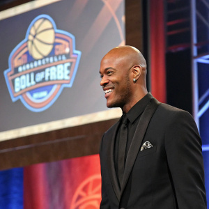 Stephon Marbury, an ex-NBA player, at a basketball hall of fame induction ceremony. He is wearing a black suit, bald head, and a confident smile. The backdrop features a large screen displaying highlights from his career.