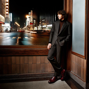 Elliot Gould, donning a black suit with maroon boots, stands against a bustling city street background, adorned with neon lights and modern billboards.