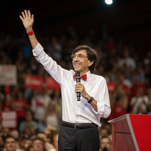 Elio Di Rupo waving enthusiastically at a political rally. Crowd in the background, blurred
