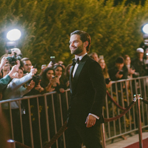 Tobey Maguire at an awards show, wearing a black tuxedo. Surrounded by flashing cameras and crowds of people.
