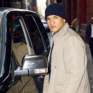 Nathaniel Arcand, starring as Clinton Skye in 'FBI Most Wanted', stands beside a sleek black SUV. The actor wears a dark beanie and a high-collared jacket. The backdrop is a dimly lit city street, with blurred figures in the background.
