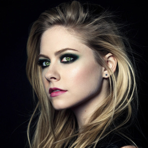 Promotional image for Avril Lavigne. Close-up of her face with striking green eyes and bold makeup. The black background emphasizes her makeup and features. Dramatic lighting highlights her beauty.