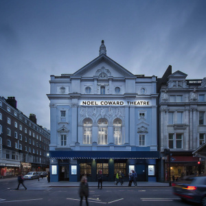 A bustling street scene in London. Ornate facade of the Noël Coward Theatre stands out, adorned with intricate details and a large sign.