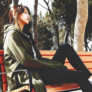 Yoona, Asian actress, dressed casually in a green hoodie and black leggings, sitting on a bench. Outdoor park setting, trees in background.