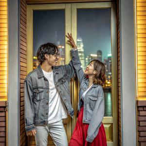 Pakho Chau and Charlene Choi at the promotional event for the film 77 Heartbreaks. The backdrop is a vibrant city skyline. Both actors are dressed casually. Pakho in a denim jacket and white shirt, Charlene in a denim jacket and red skirt.