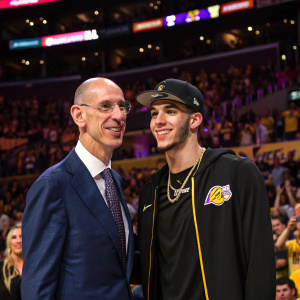 Adam Silver and Lonzo Ball at the Lakers game opening night. Silver in a navy suit, Lonzo in a black jersey and cap. The crowd cheers in the background.