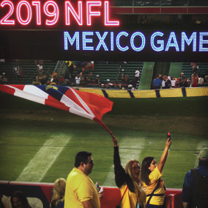Throngs of diverse fans cheering at 2019 NFL International Series match in Mexico City. A massive banner reading '2019 NFL Mexico Game' in vibrant blue letters is prominently displayed.