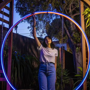 Photographic portrait of Phoebe Cates, dressed in a casual white t-shirt and jeans. Poses in a suburban backyard, surrounded by greenery.