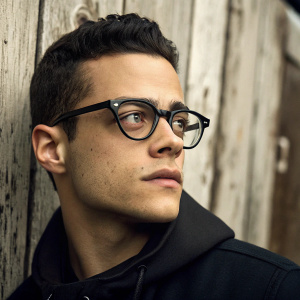 Close-up portrait of Rami Malek from Mr. Robot. Serious expression, thick black frame. Rustic, textured wooden wall background.