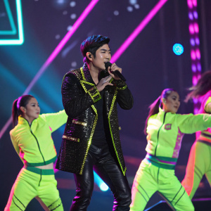 Jay Chou, a renowned performer, on stage at the Grammy Awards. Surrounded by dancers in neon outfits. Festive atmosphere with bright lights
