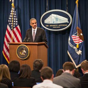 Press conference featuring Eric Holder, former Attorney General. Formal attire, podium setup. Flag and emblematic backdrop.