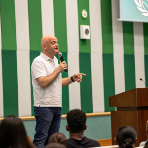 Gianni Infantino in casual attire giving a speech at the UN. Backdrop is green and white striped wall. Audience is visible.