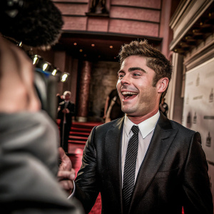 Zac Efron at a movie premiere. Wearing black suit and tie. On red carpet with repeated logos backdrop.