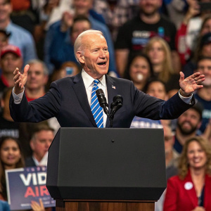 Joe Biden, former US President, addressing a rally. The venue is packed with supporters. Wearing a dark suit and a blue striped tie, he's animatedly gesturing with his hands.