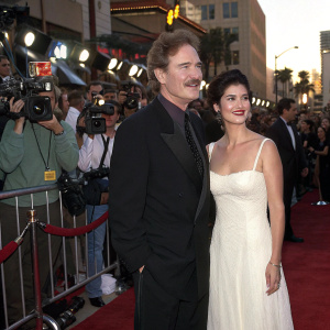 Kevin Kline and Phoebe Cates at a bustling Hollywood premiere. Kevin, dressed in a sleek black suit, Phoebe in a stunning white gown. Red carpet scene with flashing cameras and excited fans.