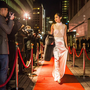 Araya Hargate at the Toronto International Film Festival. She wears a stunning white silk gown adorned with delicate lace detailing. The gown has a high neckline and flowing skirt. A fur stole in a contrasting shade adds a touch of elegance. Red carpet with paparazzi in the background.