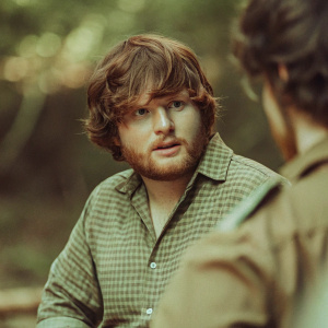 Ed Sheeran, in a candid moment. Big beard, tousled brown hair. Wearing a casual checkered shirt. Dark background highlighting his face. High-quality, professional photo.