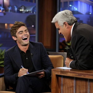 Zac Efron laughing heartily, reacting to Jay Leno's joke on The Tonight Show set.