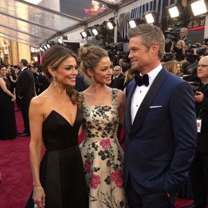 Maria Menounos, Rebecca Gayheart, and Eric Dane attending the Oscars. Maria in a black evening gown, Rebecca in a floral print dress, Eric in a navy blue tuxedo. Crowded red carpet scene with paparazzi
