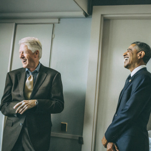 Bill Clinton and Barack Obama laughing together. Light blue background, formal attire.