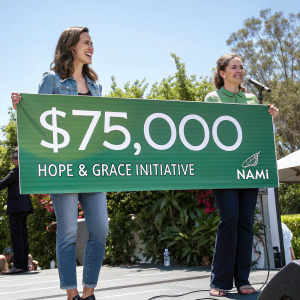 Torrey DeVitto and Mary Giliberti standing on a sunny outdoor stage. They are holding a large green banner announcing a $75,000 donation from the Hope & Grace Initiative to NAMI.