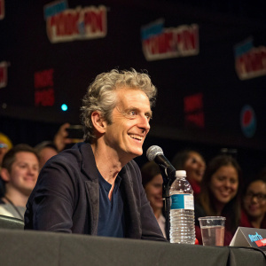 Peter Capaldi during a panel discussion at New York Comic Con. Sitting at a table with a microphone, water bottle, and glass. Crowd of excited fans in the background.