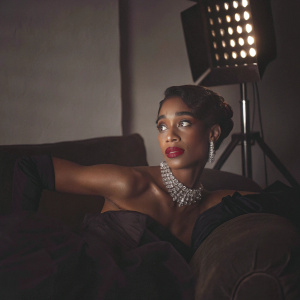Laura Harrier, elegant and confident, posing for a magazine cover shoot. She wears a glamorous black dress, adorned with a silver necklace and statement earrings. The backdrop is a plain white wall, spotlighting her striking features and bold makeup.