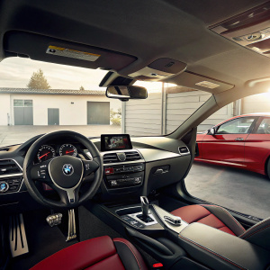 Interior of a 2014 BMW M235i Coupe. Sleek dashboard, digital display, and steering wheel with BMW emblem. Parked in garage beside red car.