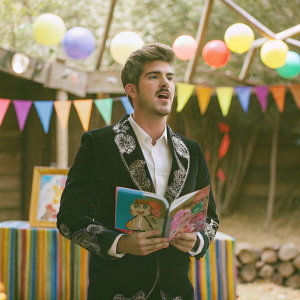 Joey Graceffa at the Children's EDEN book launch party, wearing a black velvet blazer with silver embroidery, paired with a crisp white shirt. He holds a vibrant Children's EDEN book. The background features a colorful stage setup.