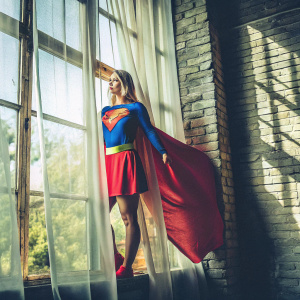 Melissa Benoist, dressed as Supergirl, standing in a room with brick walls and sheer curtains. The room is filled with soft natural light. She wears a superhero costume with the Superman emblem and a red skirt.