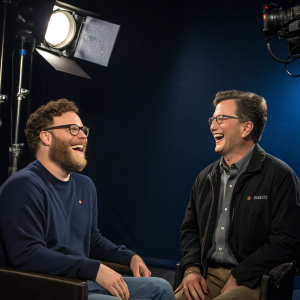 Seth Rogen and Stephen Colbert laughing off-camera during a commercial break at Comedy Central. Studio lights, dark blue backdrop.