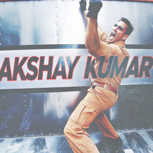Akshay Kumar in a dynamic action pose, wearing an orange jumpsuit and khaki uniform. Promotional backdrop for his upcoming film.