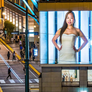 Professional headshot of a young Asian woman with long brown hair. She is standing confidently in a light-colored, strapless dress against a busy street backdrop with people walking by.