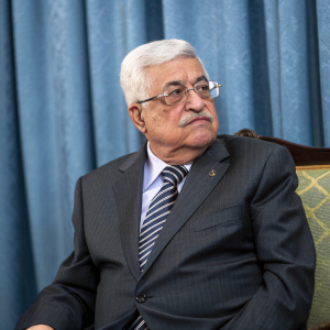 Mahmoud Abbas, the Palestinian leader, in a stern pose. Dressed in a dark grey suit and striped tie. Blue curtain backdrop.