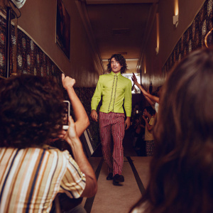 Ezra Miller in a candid shot at Comic-Con. He is wearing a neon shirt. Background is a sea of fans and event banners.