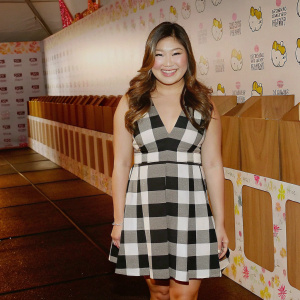Jenna Ushkowitz, with a warm smile, dressed in a black and white plaid dress, stands at a Hello Kitty themed charity event. Brightly colored backdrop with Hello Kitty logos and sponsors