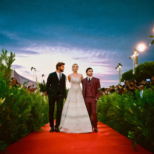 Liam Hemsworth, Jennifer Lawrence, and Josh Hutcherson on the red carpet for The Hunger Games premiere. Liam in black suit, Jennifer in white detailed gown, Josh in maroon suit. Crowded backdrop with lights and paparazzi.