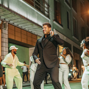 Michael Bublé in a vibrant music video. He's wearing a sharp black suit. The backdrop is a bustling city street at night. Various people in casual outfits are dancing and enjoying themselves.