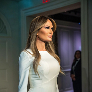 A wax figure of Melania Trump. Pale skin, long flowing hair. Wearing a white dress. Stands in front of a dimly lit room with subtle shades