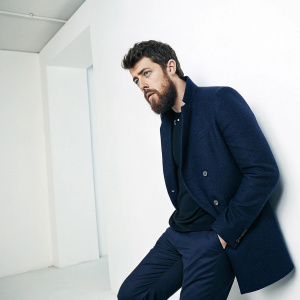 Toby Kebbell, bearded and sharp in a navy blazer, poses solo in a stark white studio setting.
