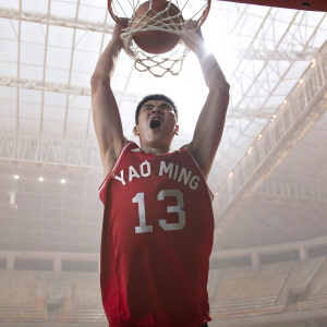 Yao Ming, with a fierce look, mid-air during a slam dunk. His red jersey with the number 13 stands out. The arena is empty, spotlight on
