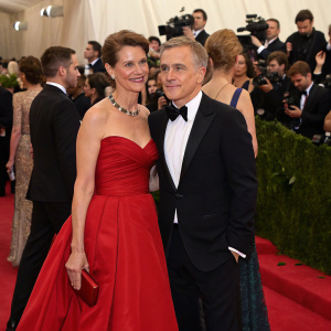 Judith Holste and Christoph Waltz at the Met Gala. Judith in a stunning red gown, Christoph in a black tuxedo. Crowded event, flash photography.