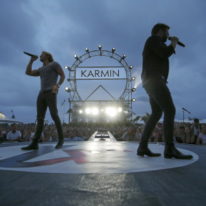 Karmin band members performing on stage. Illuminated backdrop with band logo. Crowd in background