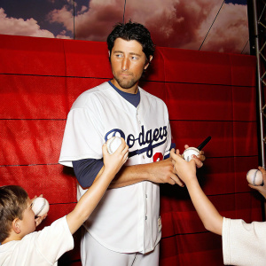 Nomar Garciaparra signing autographs in a white LA Dodgers jersey. Baseballs and outstretched hands surround him. Cloudy stadium backdrop