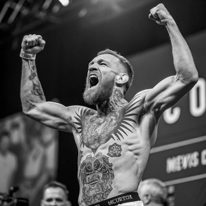 Conor McGregor, the UFC champion, in a stark black and white photo. Celebrating victory with flexed muscles and a fierce expression. Intricate tattoos on his torso. Blurred crowd and stage backdrop.