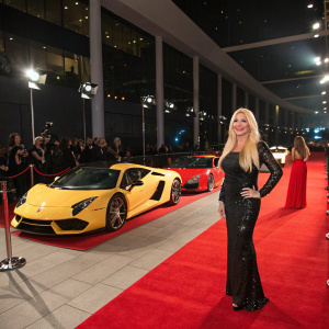 Monika Bacardi at a luxury car unveiling event, smiling in a red carpet attire. Surrounded by Lamborghini sports cars