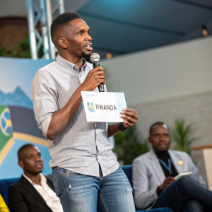 Samuel Eto'o, former football star, in a casual shirt and jeans, speaking at a charity event. Holding a sign that says 'Rwanda'.